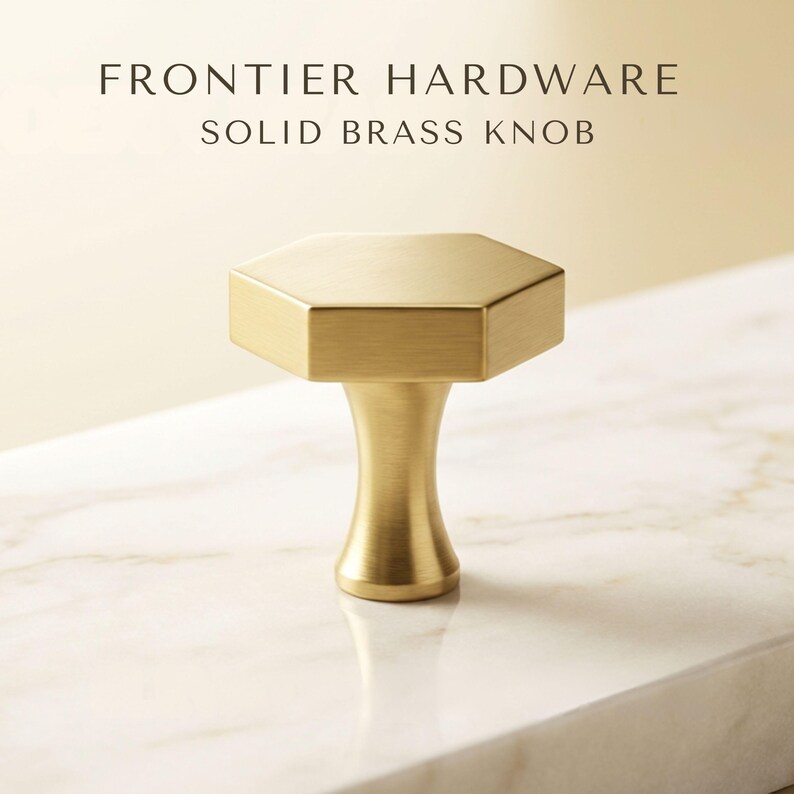 May include: A solid brass knob with a hexagonal top and a tapered base. The text FRONTIER HARDWARE SOLID BRASS KNOB is displayed above the object. The knob has a brushed metallic finish and a distinct geometric shape with a smooth, flared stem.