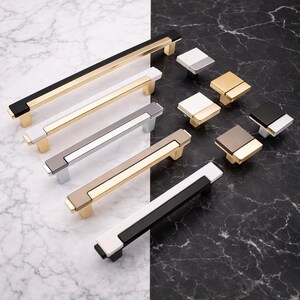 Peut inclure: Une collection de quincaillerie de meubles moderne, comprenant des barres de tirage et des boutons carr&eacute;s. Les poign&eacute;es ont un design rectangulaire avec des accents dor&eacute;s, noirs, blancs, argent&eacute;s et taupe. Les boutons sont carr&eacute;s avec des couleurs similaires.
