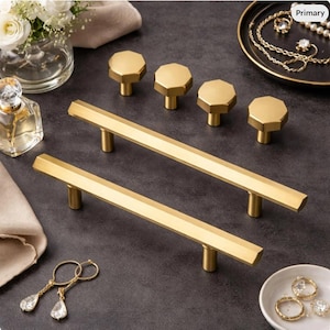 May include: A set of gold-toned cabinet hardware including two long, linear drawer pulls with a faceted, hexagonal profile and four matching hexagonal knobs, all featuring a smooth, metallic finish and cylindrical mounting posts.