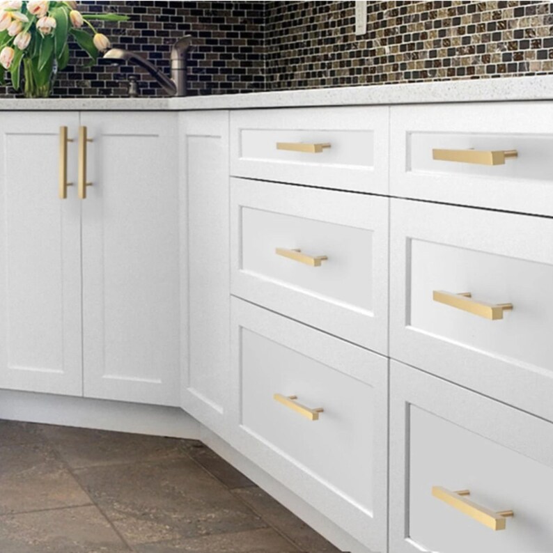 Pode incluir: Um conjunto de arm&aacute;rios e gavetas de cozinha brancos com puxadores de barra em metal dourado escovado, dispostos horizontal e verticalmente. Os m&oacute;veis possuem um design de painel plano com acabamento liso e visual luminoso.