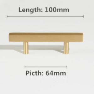 Pode incluir: Um puxador de m&oacute;vel em metal dourado com uma barra retangular e dois postes de montagem cil&iacute;ndricos. O texto indica um comprimento total de 100 mm e um espa&ccedil;amento entre furos, rotulado como "Picth", de 64 mm. A pe&ccedil;a tem um acabamento mate liso.
