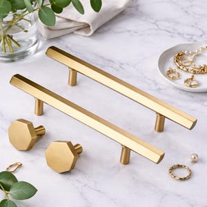 May include: Two long, gold-toned metal cabinet pulls with a hexagonal profile and two matching octagonal cabinet knobs. The hardware features a smooth, brushed finish and a geometric design, presented as a set of four individual furniture components.