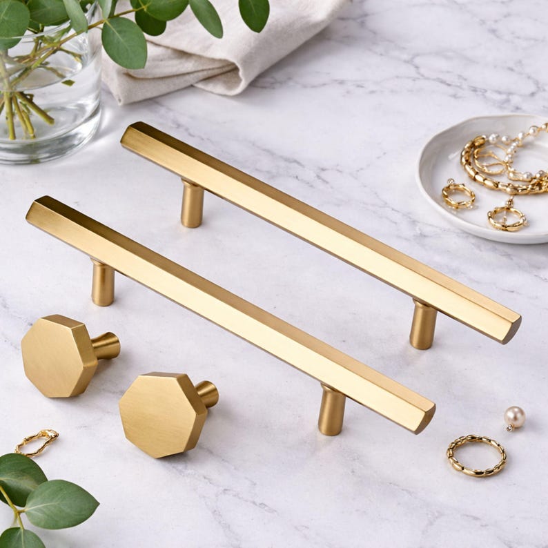 May include: Two long, gold-toned metal cabinet pulls with a hexagonal profile and two matching octagonal cabinet knobs. The hardware features a smooth, brushed finish and a geometric design, presented as a set of four individual furniture components.