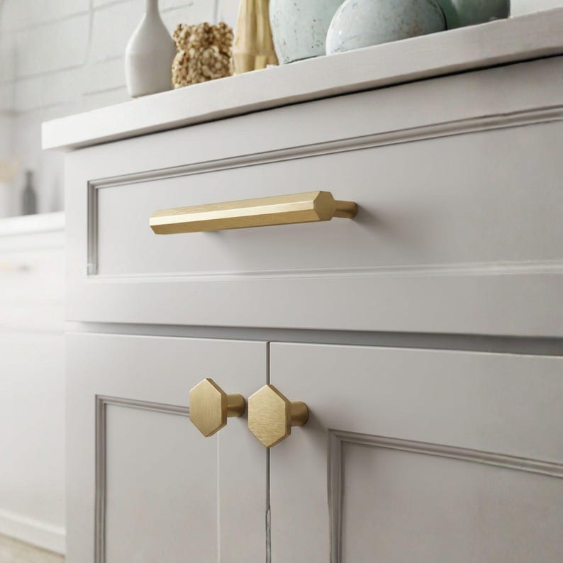 May include: A brushed gold cabinet handle with a long hexagonal bar shape and two matching hexagonal knobs installed on light gray shaker-style cabinet doors. The hardware features a matte metallic finish and geometric, multi-faceted edges.