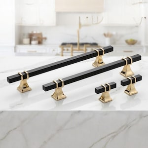 May include: A set of black and gold cabinet hardware. The hardware includes two long rectangular pulls, and four smaller square knobs. The pulls and knobs have a black, rectangular bar with gold-colored accents and bases.