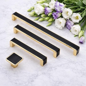 May include: A set of gold-toned cabinet hardware with black accents. The set includes three rectangular handles of varying lengths and a square knob. White and purple flowers are in the background.