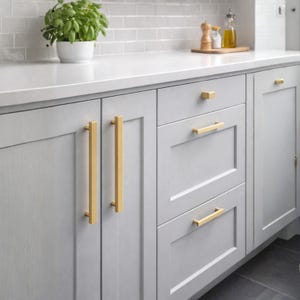 Pode incluir: Arm&aacute;rios de cozinha cinza-claros com puxadores de barra longos e verticais em metal dourado escovado nas portas e puxadores de barra horizontais dourados nas gavetas. Os arm&aacute;rios t&ecirc;m um design de painel plano com acabamento fosco.