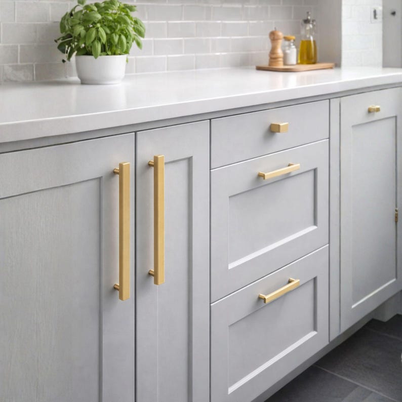 Pode incluir: Arm&aacute;rios de cozinha cinza-claros com puxadores de barra longos e verticais em metal dourado escovado nas portas e puxadores de barra horizontais dourados nas gavetas. Os arm&aacute;rios t&ecirc;m um design de painel plano com acabamento fosco.