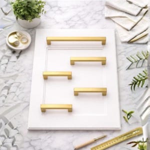 May include: A white rectangular cabinet door panel featuring five gold-toned metal drawer pulls of varying lengths arranged in a staggered pattern. The hardware has a smooth, rectangular profile and is mounted directly onto the flat surface.