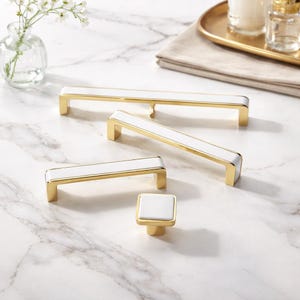 May include: A collection of cabinet hardware including three rectangular drawer pulls of varying lengths and one square cabinet knob. Each piece features a polished gold metal frame surrounding a smooth, solid white rectangular or square center inlay.