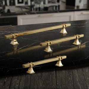 May include: Four gold-colored cabinet pulls of varying sizes are displayed on a black marble countertop. Each pull features a rectangular bar with decorative square accents and a square base. The pulls are designed for kitchen or bathroom cabinetry.