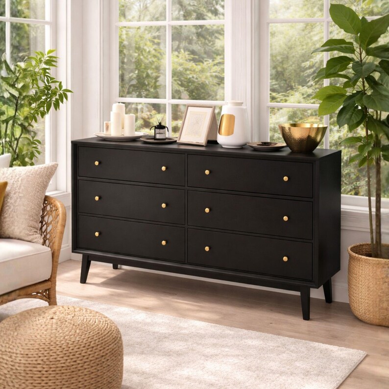 May include: A black six-drawer dresser featuring gold-toned circular knobs. The dresser is supported by four tapered legs and has a rectangular frame. On top of the dresser are several white candles, a small framed picture, a white vase, and a gold bowl.