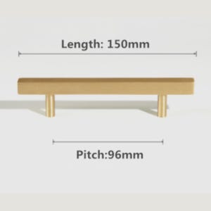 Pode incluir: Um puxador de m&oacute;vel em metal dourado com uma barra retangular e dois postes de montagem cil&iacute;ndricos. O texto indica um comprimento total de 150mm e uma dist&acirc;ncia entre furos de 96mm. A ferragem possui um acabamento mate e liso.
