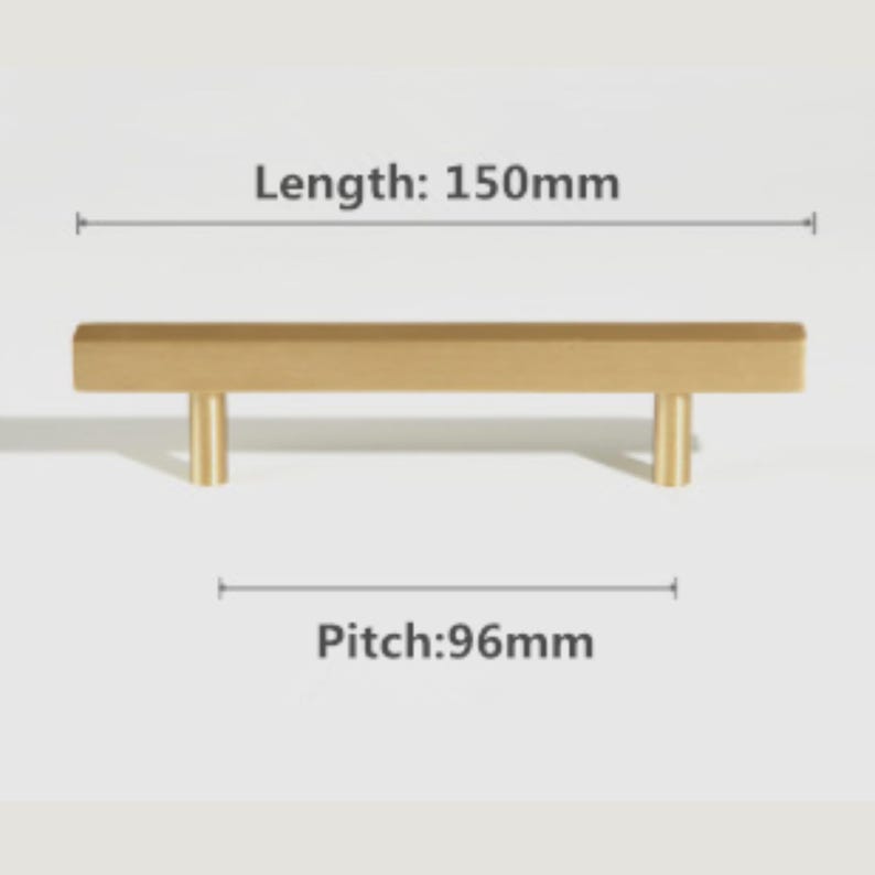 Pode incluir: Um puxador de m&oacute;vel em metal dourado com uma barra retangular e dois postes de montagem cil&iacute;ndricos. O texto indica um comprimento total de 150mm e uma dist&acirc;ncia entre furos de 96mm. A ferragem possui um acabamento mate e liso.