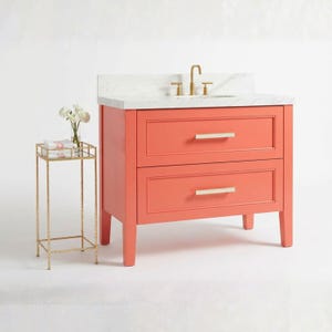 May include: A coral-colored bathroom vanity with two drawers and a white marble countertop. The vanity has gold-toned hardware and a matching gold side table with a floral arrangement. The faucet is gold-toned.