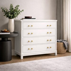 May include: A white four-drawer dresser featuring gold-toned circular knobs on each drawer. The dresser stands on a flat base and is positioned next to a black cylindrical side table holding a white vase with green foliage and a stack of books.