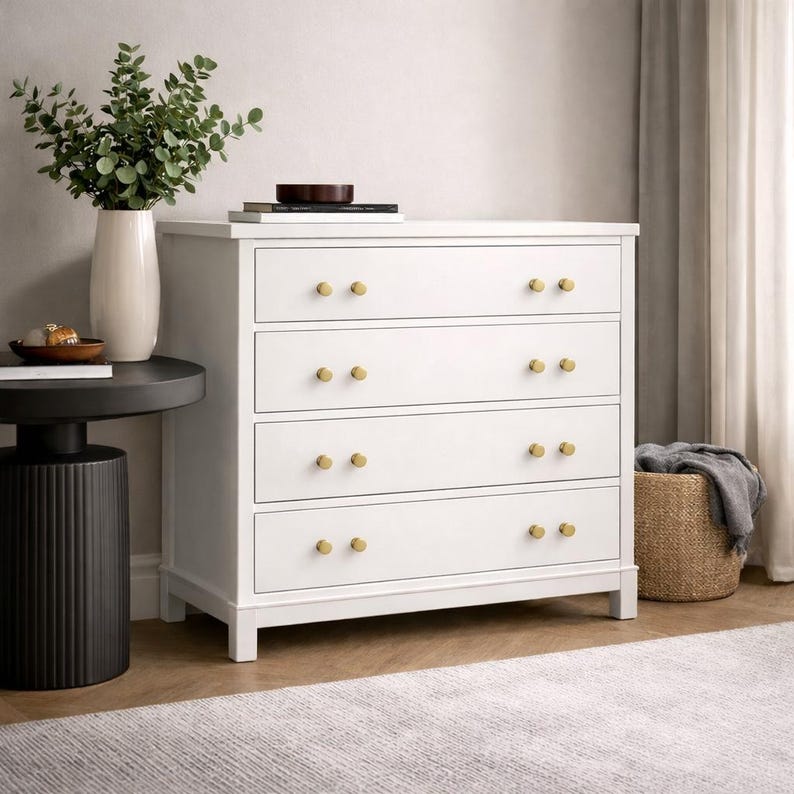 May include: A white four-drawer dresser featuring gold-toned circular knobs on each drawer. The dresser stands on a flat base and is positioned next to a black cylindrical side table holding a white vase with green foliage and a stack of books.