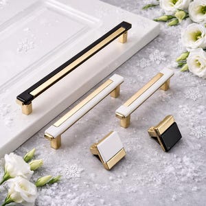 May include: A collection of cabinet hardware, including handles and knobs. The handles feature a gold center stripe with black or white accents. The knobs have a square design with gold and black or white finishes. The hardware is displayed on a white surface.