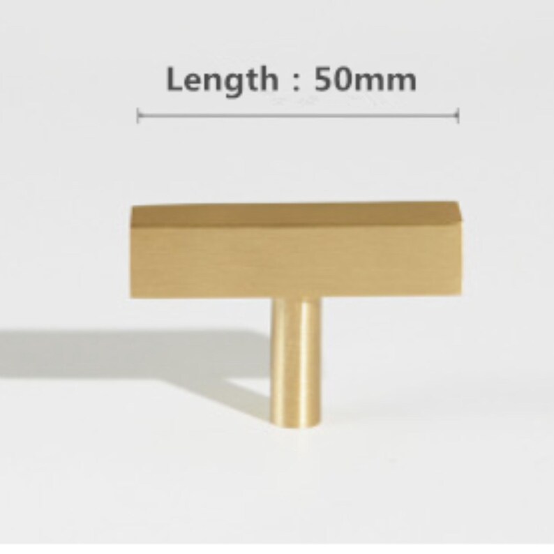 Pode incluir: Um puxador de m&oacute;vel em forma de T de cor dourada com acabamento met&aacute;lico escovado. O puxador apresenta uma barra retangular horizontal fixada a uma base cil&iacute;ndrica. O texto indica um comprimento de 50mm com uma linha de medi&ccedil;&atilde;o.