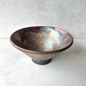 May include: A handcrafted, iridescent ceramic bowl with a wide, shallow design. The bowl features a metallic finish with hues of bronze, gold, and hints of blue and purple. It sits on a dark gray, circular base, creating a striking contrast.