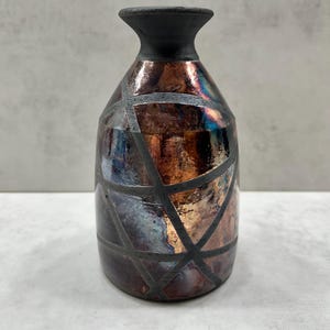 May include: A small vase with a narrow neck and a wide body, featuring a metallic, iridescent finish in shades of copper, bronze, and blue. The surface is decorated with a geometric pattern of intersecting matte black lines across the body.