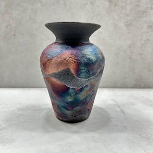 May include: A small vase with a flared rim and a dark gray matte neck. The body features a textured, swirling finish with metallic hues of copper, blue, and purple, accented by a band of rough, dark gray speckled material across the center.