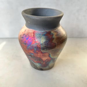 May include: A small, handmade ceramic vase with a dark gray rim and a body with iridescent colors of red, blue, and gold. The vase has a rounded shape and a narrow neck, suitable for displaying small flowers or as a decorative piece.