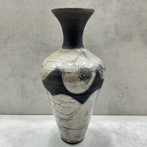 May include: A ceramic vase featuring a flared rim and a narrow neck in matte black. The body displays a white crackle glaze finish with dark, web-like fissures and bold, black geometric shapes encircling the midsection of the vessel.