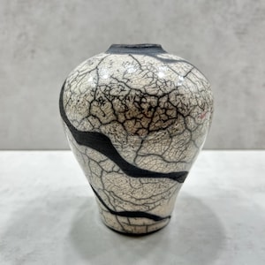 May include: A ceramic vase with a rounded body and a narrow opening, featuring a cream-colored surface covered in a dense network of fine black crackle lines and bold, smooth black horizontal bands that wrap around the exterior of the vessel.