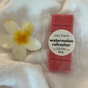May include: A red wax snapbar in clear plastic packaging with a circular label reading Juicy Guava watermelon refresher Soy Wax Snapbar hand poured in Vancouver, CAN 55 g, accompanied by a white and yellow plumeria flower hair claw clip.