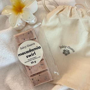 Peut inclure: Une tablette de cire de soja de 55 g dans un emballage plastique transparent avec une étiquette Juicy Guava macadamia swirl, accompagnée d'une pochette beige à cordon avec un logo hibiscus et une pince à cheveux fleur blanche et jaune.