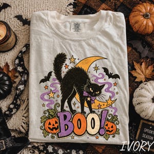 May include: An ivory t-shirt featuring a graphic of a black cat with a purple polka dot bow, standing over the word BOO in large letters. The design includes a yellow crescent moon, orange jack-o'-lanterns, purple smoke, and stars.