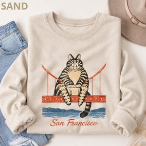 May include: A sand-colored crewneck sweatshirt featuring a graphic of a tabby cat sitting on the Golden Gate Bridge. Below the bridge is a blue water illustration and the text San Francisco in a red script font. The sweatshirt has long sleeves.