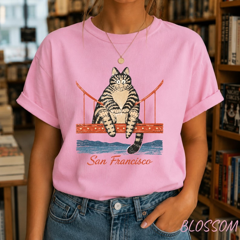 May include: A light pink short-sleeve t-shirt featuring a graphic of a tabby cat sitting on the Golden Gate Bridge. Below the bridge, the text San Francisco is printed in a cursive font. The cat has wide eyes and a tan belly.
