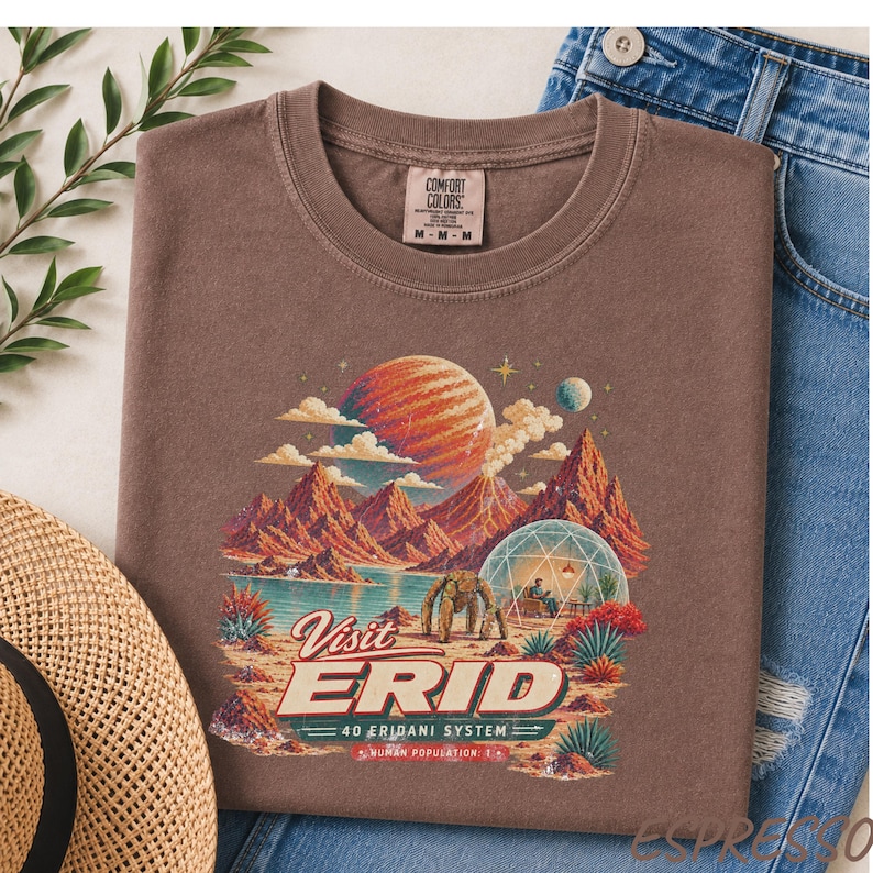 May include: A brown t-shirt featuring a graphic print of a space landscape with a large planet, mountains, a geodesic dome, and a creature. The text reads Visit ERID 40 ERIDANI SYSTEM HUMAN POPULATION: 1. The shirt has a Comfort Colors brand label.