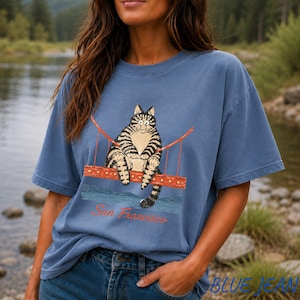 May include: A dusty blue short-sleeve t-shirt featuring a graphic of a brown tabby cat sitting on a red suspension bridge. Below the bridge, the text San Francisco is printed in a cursive red font. The shirt has a crew neckline and a relaxed fit.