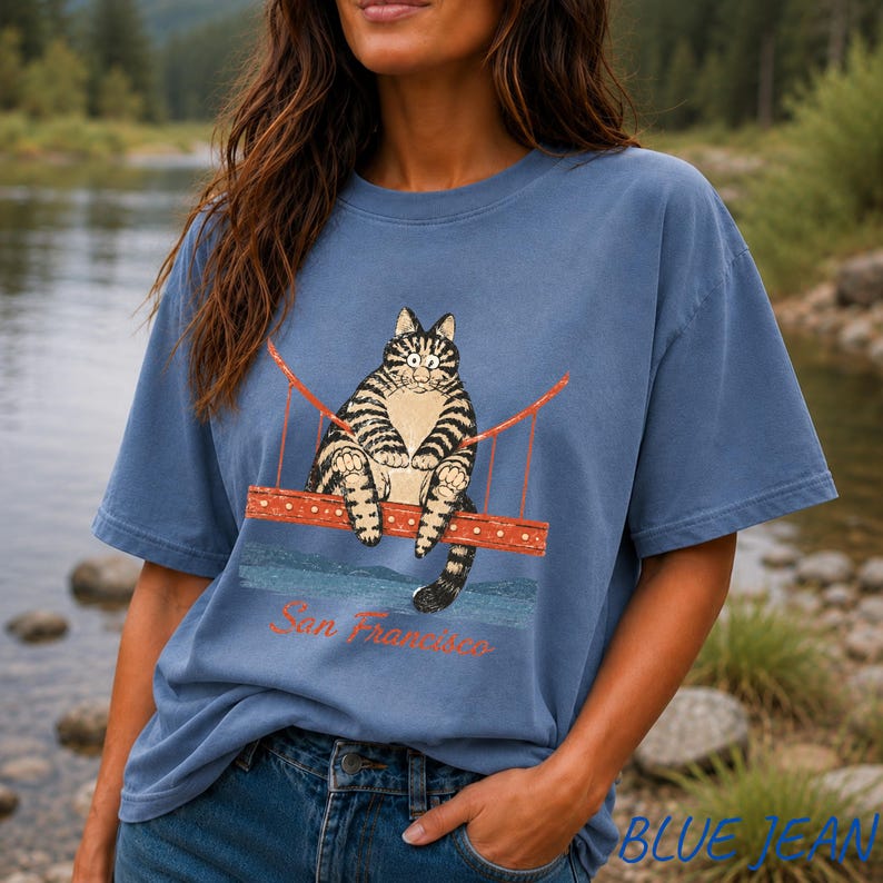 May include: A dusty blue short-sleeve t-shirt featuring a graphic of a brown tabby cat sitting on a red suspension bridge. Below the bridge, the text San Francisco is printed in a cursive red font. The shirt has a crew neckline and a relaxed fit.