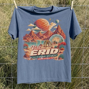 May include: A dusty blue short-sleeve t-shirt featuring a retro-style space travel graphic. The print depicts a red planet, mountains, a geodesic dome, and a robot, with the text "Visit ERID 40 ERIDANI SYSTEM HUMAN POPULATION 1" in cream and red.