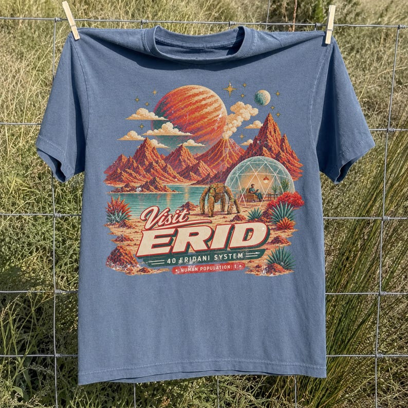 May include: A dusty blue short-sleeve t-shirt featuring a retro-style space travel graphic. The print depicts a red planet, mountains, a geodesic dome, and a robot, with the text "Visit ERID 40 ERIDANI SYSTEM HUMAN POPULATION 1" in cream and red.