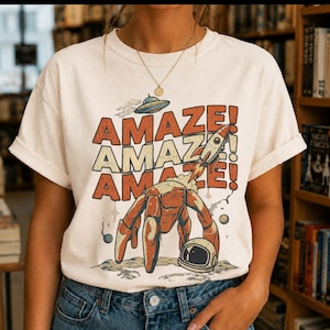May include: A cream-colored short-sleeve t-shirt featuring a graphic print with the text AMAZE! repeated three times in orange. The design includes a brown crab, a rocket ship, a flying saucer, and an astronaut helmet on a lunar surface.