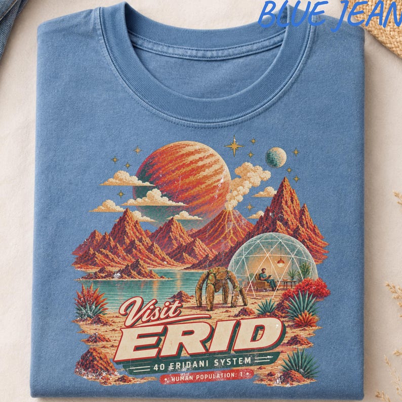 May include: A blue t-shirt featuring a graphic print of a space landscape with a large orange planet, red mountains, a geodesic dome, and a robotic figure. The text reads: Visit ERID, 40 ERIDANI SYSTEM, HUMAN POPULATION: 1.