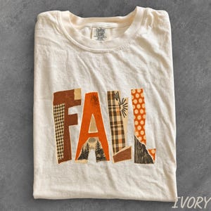 May include: An ivory short-sleeve t-shirt featuring the word FALL printed in large, collage-style letters. Each letter displays a different pattern, including houndstooth, solid orange, plaid, and a forest scene with trees and a sun.