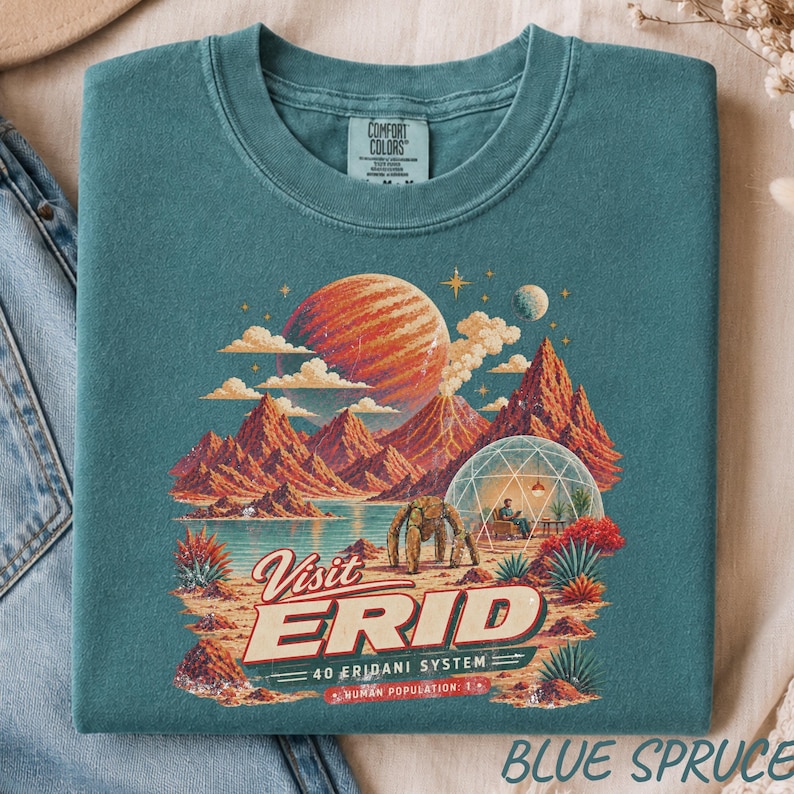 May include: A blue-green crewneck t-shirt featuring a graphic of a space landscape with a large planet, mountains, a dome structure, and a robotic figure. The text reads Visit ERID 40 ERIDANI SYSTEM HUMAN POPULATION: 1 in a retro font style.