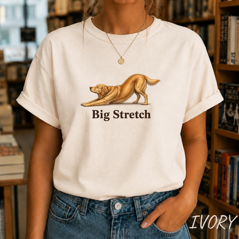 May include: An ivory-colored short-sleeve t-shirt featuring a graphic illustration of a golden-colored dog in a stretching pose. Below the dog, the text "Big Stretch" is printed in a dark brown, serif font. The shirt has a crew neckline.