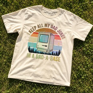 May include: A cream-coloured short-sleeve t-shirt featuring a graphic of a vintage computer. The design includes a sunset-coloured circle, a silhouette of pine trees, and the text: I KEEP ALL MY DAD JOKES IN A DAD-A-BASE, printed in a distressed font.