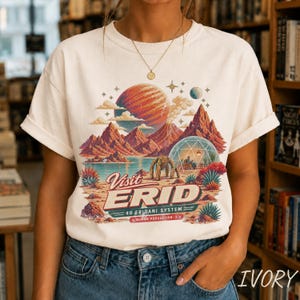May include: An off-white t-shirt featuring a colorful graphic of a red planet, mountains, a geodesic dome, and a robotic figure. The text reads Visit ERID 40 ERIDANI SYSTEM HUMAN POPULATION: 1. The design includes desert plants and clouds.