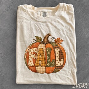 May include: An ivory t-shirt featuring a large graphic of a pumpkin with the word FALL spelled out across its segments. The pumpkin design incorporates various patterns including floral, plaid, and polka dots in shades of orange, brown, and green.
