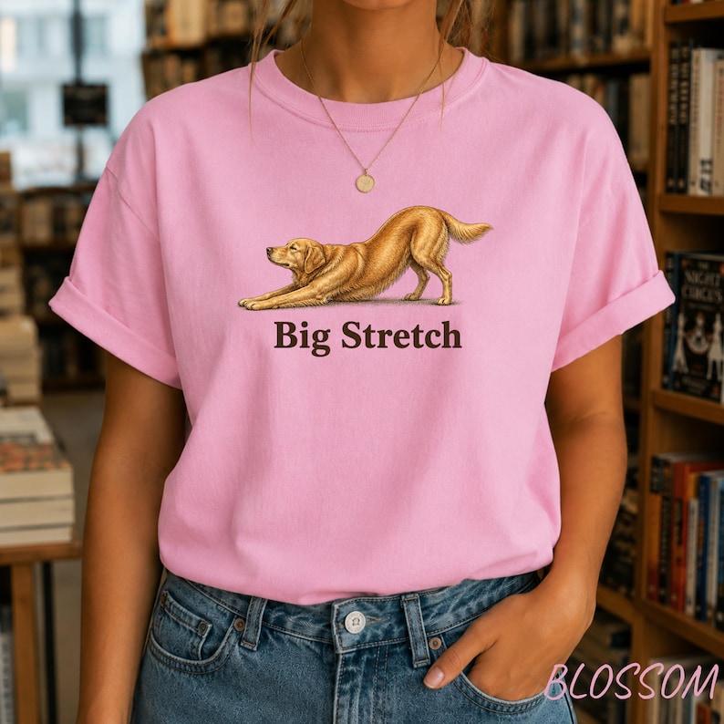 May include: A light pink short-sleeve t-shirt featuring a graphic illustration of a golden-colored dog in a stretching pose. Below the dog, the text "Big Stretch" is printed in a dark brown font. The shirt has a crew neckline and rolled sleeves.
