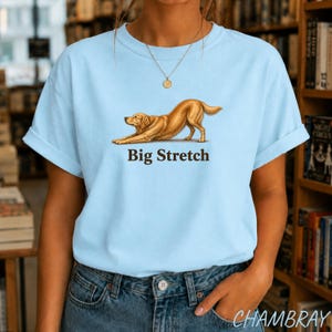 May include: A light blue t-shirt featuring a graphic of a golden-colored dog in a stretching pose. Below the dog, the text "Big Stretch" is printed in a dark brown font. The shirt has short sleeves and a crew neckline, worn with blue denim jeans.