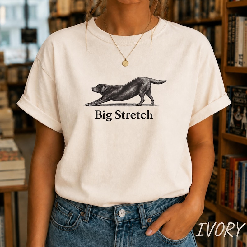 K&ouml;nnte beinhalten: Ein cremefarbenes Kurzarm-T-Shirt mit einer schwarzen Tusche-Illustration eines Hundes in einer Dehnposition. Unter dem Hund ist der Text "Big Stretch" in einer fetten, schwarzen Serifenschrift gedruckt. Das Shirt hat einen Rundhalsausschnitt und &Auml;rmelaufschl&auml;ge.